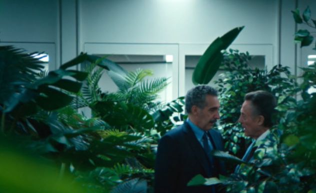 two gay office workers stand close to each other in a forest of fake plants