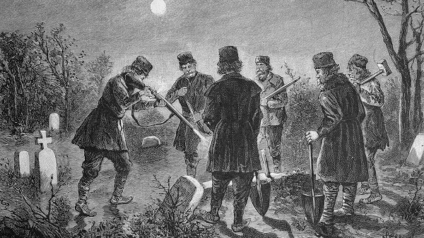 A group of men in coats and hats stand in a graveyard with shovels and watch as a man shoots a bullet into a grave
