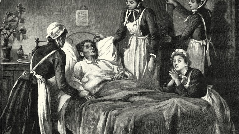 A very sick looking man lays in bed surrounded by 4 nurses who look concerned and are praying