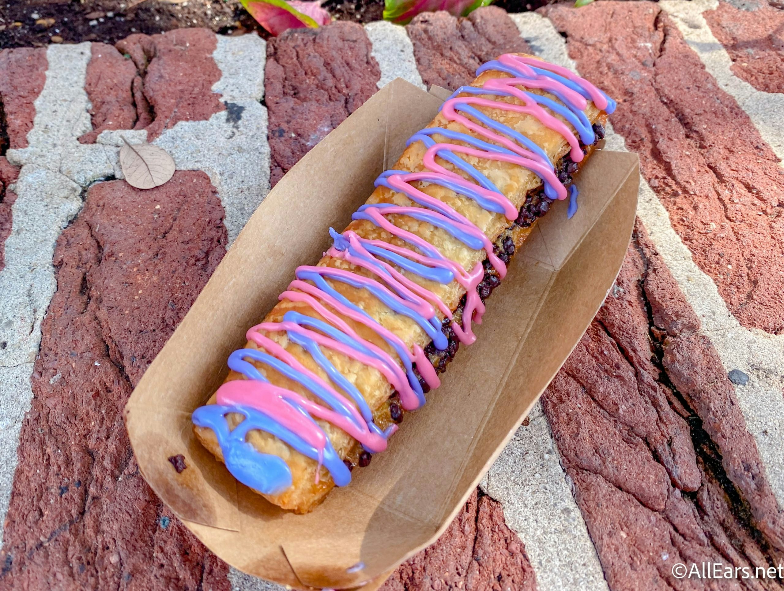 A flaky pastry sits in a candboard container, atop a brick wall. The pastry is long and crispy, with chocolate peeking out the sides, and pink and purple candy drizzle on top.