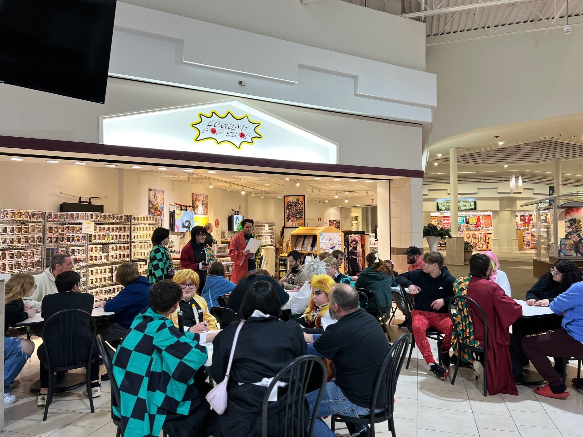 A lively gathering at a pop culture shop, with fans dressed in cosplay seated at tables participating in an event. The store’s shelves are lined with Funko Pop figures, and the scene captures a vibrant and community-oriented atmosphere.